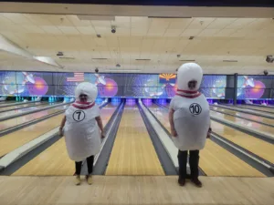 Two people dressed as number 7 and number 10 bowling pins laugh in front of several bowling lanes.