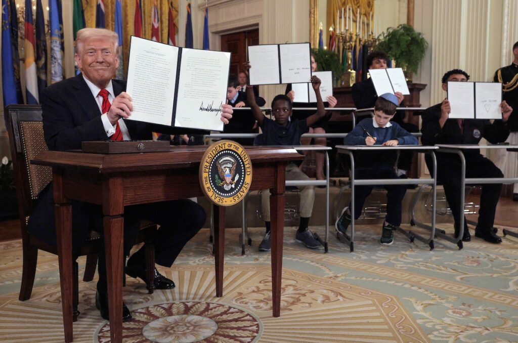 Trump sitting at desk holding up papers alongside children also sitting at desks and holding up papers